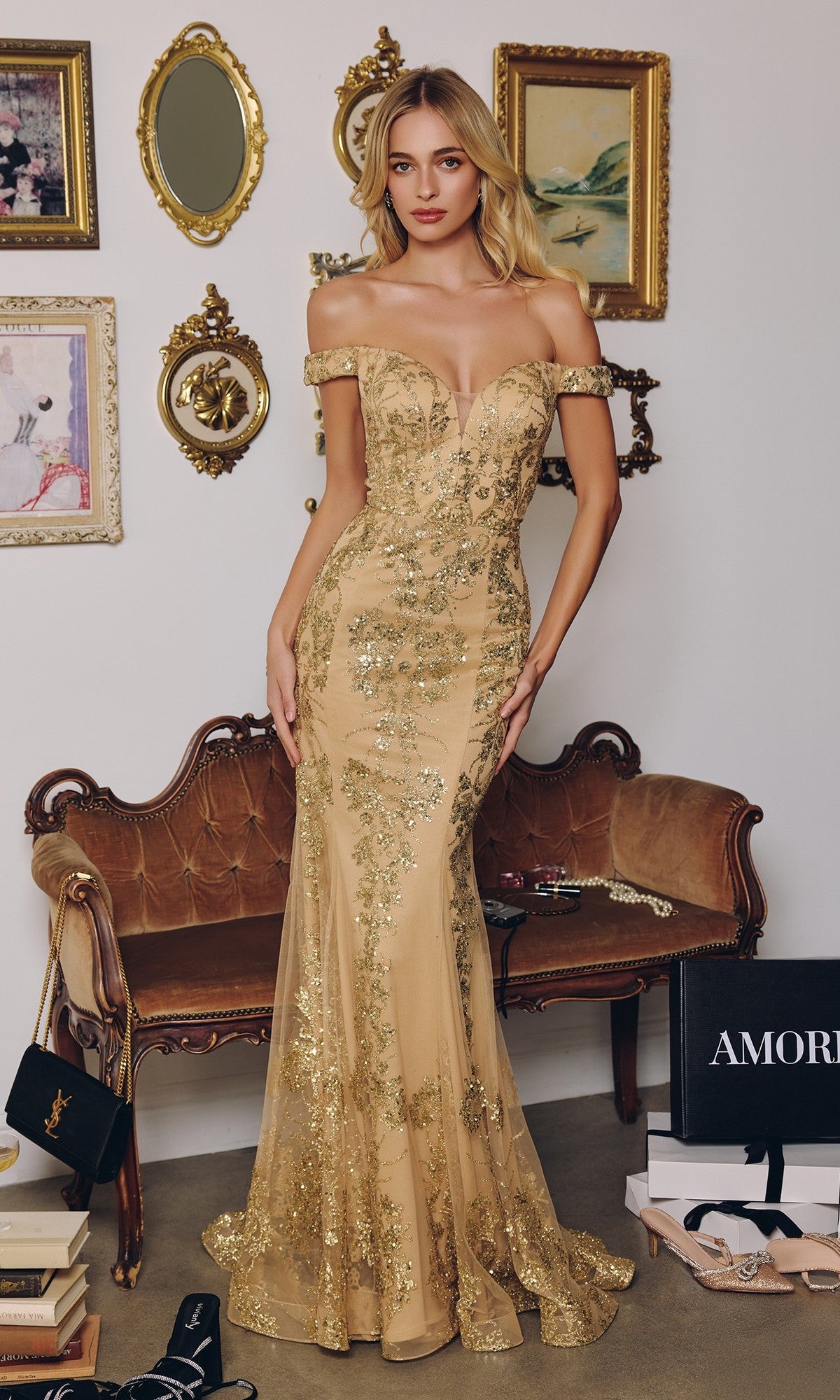 A woman stands indoors wearing the gold Off-Shoulder Sequin Prom Gown: Amoris T613. She poses in front of a brown vintage sofa, with framed art and designer shopping bags in the background.